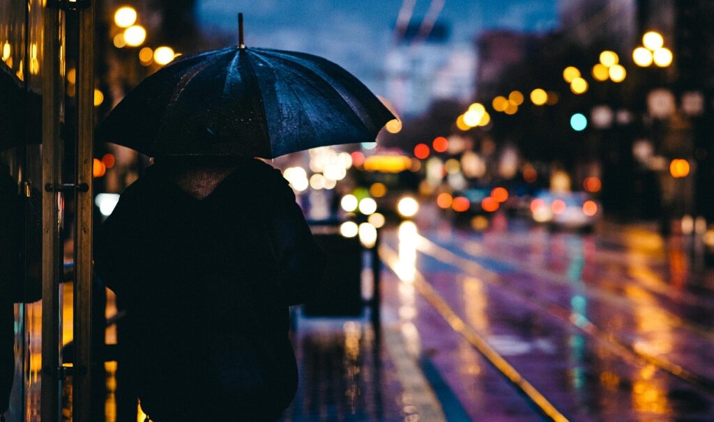 A person is seen in sharp focus holding an umbrella and wearing a jacket as rain can be seen on the streets and umbrella, with the background being blurred and full of lights from car headlights and street lights. 
