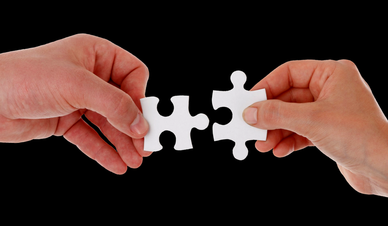 Two hands are seen holding white jigsaw pieces across from one another against a black backdrop.