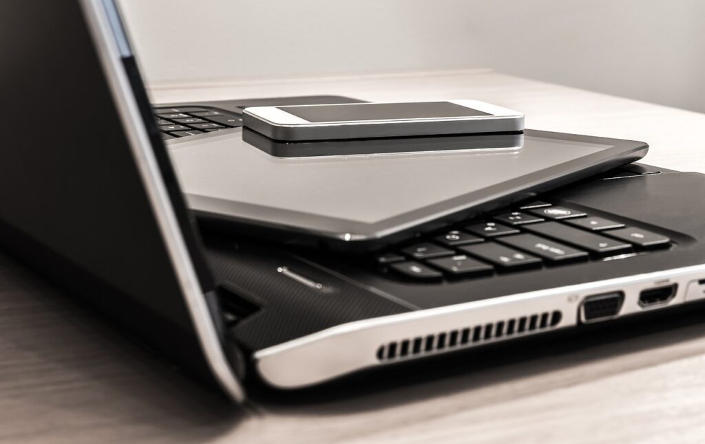 A laptop sits on a desk with a tablet device, and a smartphone piled on top of its keyboard. 