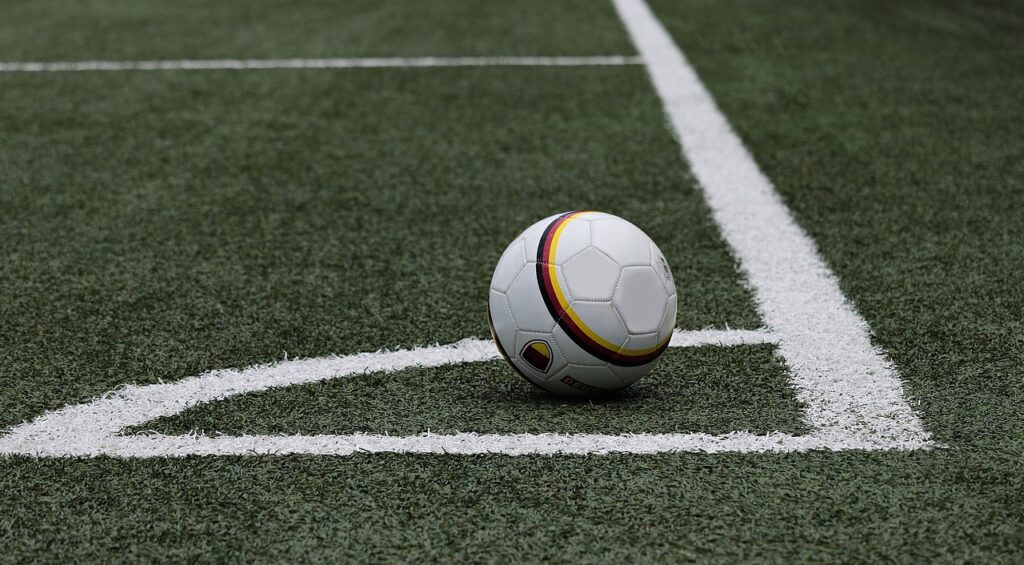 A white football with red, black and yellow accents is seen sitting in the semi-circle of the corner spot. The pitch is green in colour with white lines denoting the playing area.